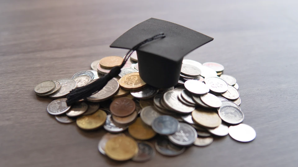 Ny låneordning for studier ved siden av jobb Bildet viser en svart akademisk hatt (mortarboard) plassert oppå en haug med mynter av forskjellige valutaer på et trebord. Dette kan symbolisere kostnadene ved utdanning eller investeringen i en akademisk fremtid.