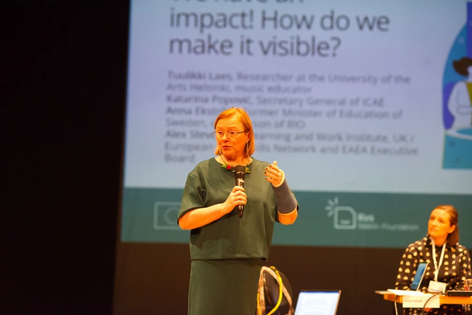 A woman is speaking on a stage, holding a microphone with her left hand. She is wearing glasses and a green outfit, and has a cast or bandage on her right arm. Behind her is a presentation slide that reads, "What an impact! How do we make it visible?" The slide also lists the names and titles of several individuals, indicating a panel or discussion event. Another woman is seated on stage, listening attentively.