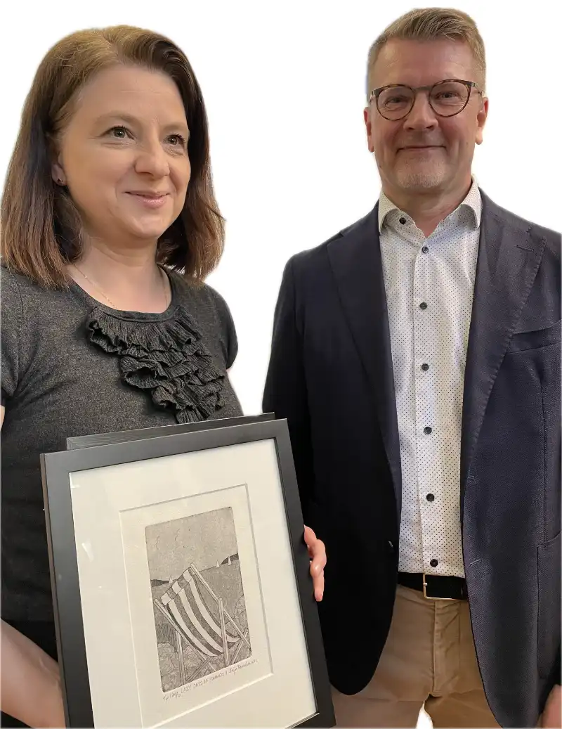 Two people are posing for a photo. The woman on the left is holding a framed artwork featuring a striped deck chair in a grassy field. She has shoulder-length brown hair and is wearing a grey top with a ruffled design. The man on the right is wearing glasses, a white shirt with small polka dots, and a dark blazer. Both are smiling and appear to be in a formal or celebratory setting.