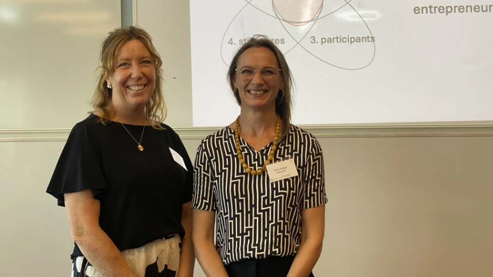 Jenny Lennhammar og Maria Kirstejn Aakjær To kvinder smilende foran en præsentationsskærm under en workshop eller konference.