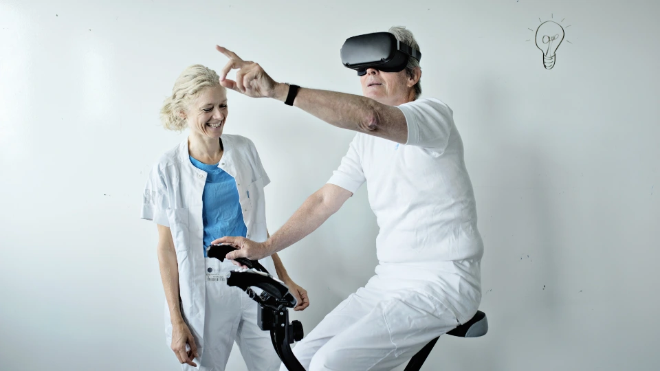 Nordisk Ministerråds politiske prioriteter 2025-2030 Ældre mand med VR-briller sidder på motionscykel og peger fremad, mens en smilende sundhedsprofessionel i hvid kittel står ved siden af. Baggrunden er en hvid væg med en tegnet pære, der symboliserer en idé.