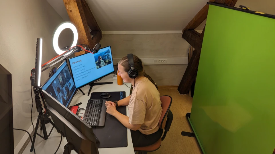 Seline Eskedal Amundsen En kvinne sitter ved en pult i et hjemmestudio med hodetelefoner på. Hun ser på to skjermer med et webmøte, omgitt av utstyr som en ringlampe og en grønn skjerm bak henne.