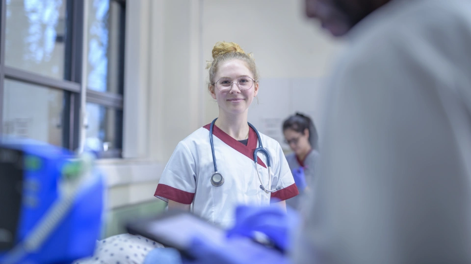En sygeplejerske i uniform med stetoskop, smilende på en hospitalsafdeling, med en kollega i baggrunden.