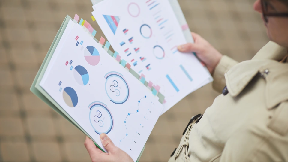 Rapport om hur den livslånga vägledningen i Finland borde utvecklas Person i beige kappa håller i dokument med färgglada diagram och grafer, organiserade med flikar.
