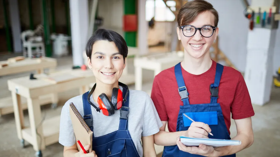 Rammer, kultur og organisering er vigtige for at sikre varig effekt af skoleudviklingsprojekter To unge håndværkere iført arbejdstøj og sikkerhedsudstyr står smilende i et træværksted med arbejdsborde og værktøj i baggrunden.