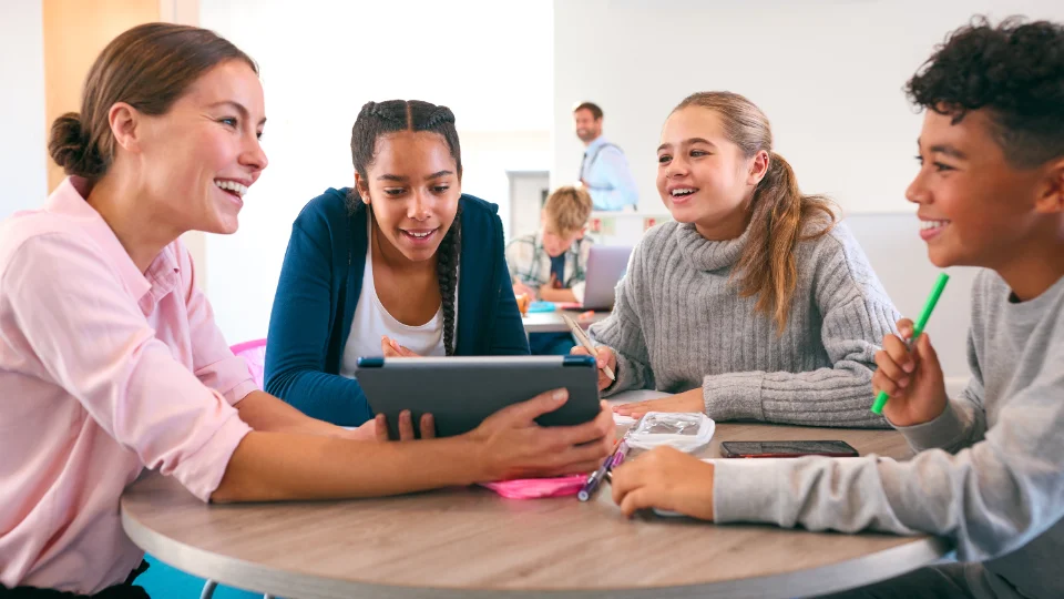 Lærer og elever sidder rundt om et bord og arbejder sammen med en tablet i et klasseværelse.