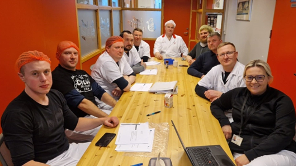 Kjøttindustrien på Island Gruppe med fabrikkarbeidere og ansatte i arbeidsklær og hårnett samlet rundt et møtebord med dokumenter og en bærbar PC.