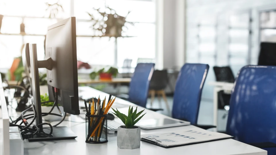 Moderne kontor med computere, blå stole, skrivebordstilbehør og grønne planter i et lyst og åbent arbejdsmiljø.