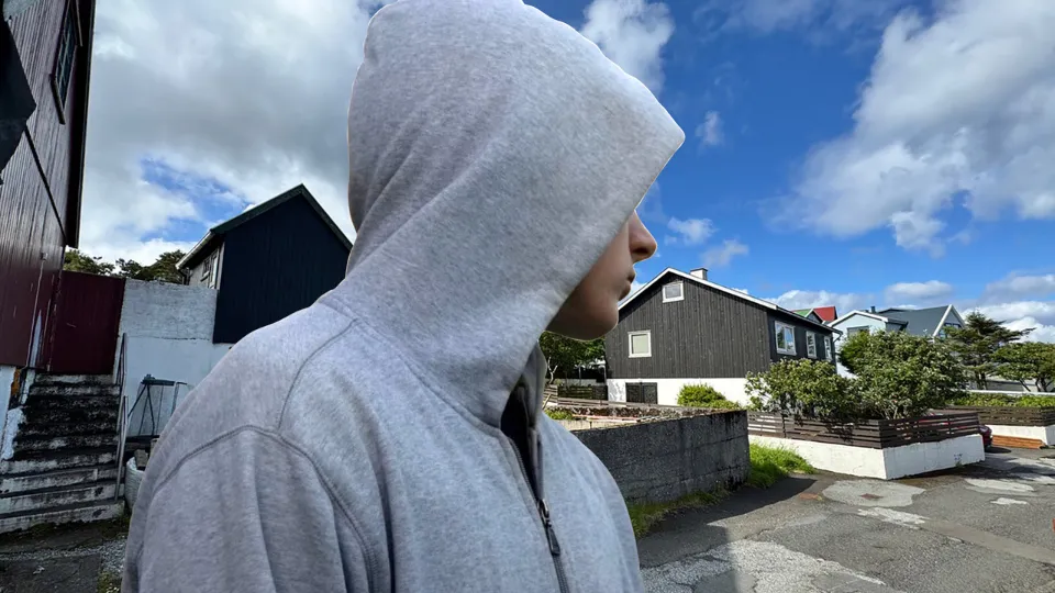 Persónur í gráum hettutrøggjum stendur úti í bygd við svørt og hvít hús undir bláum himni við skýggjum.