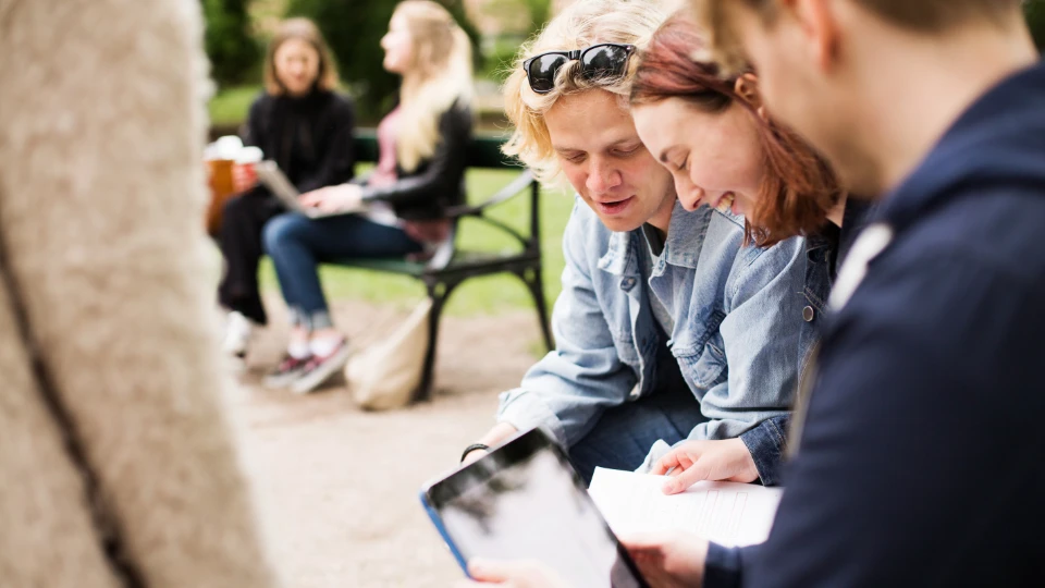 Studerende sidder udendørs i en park og samarbejder om skolearbejde med papirer og en tablet.