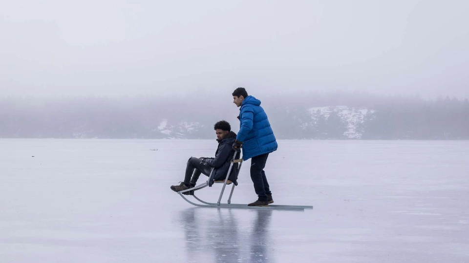 Two men on a frozen lake, one sitting on a traditional kicksled while the other pushes, surrounded by fog and winter landscape.