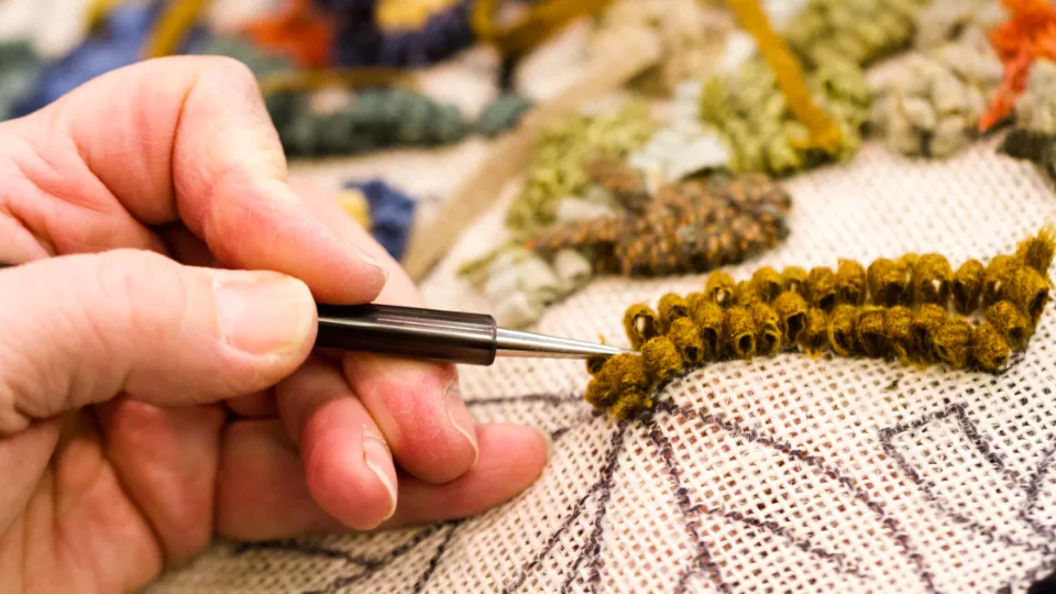 Närbild av en hand som använder en punch needle-teknik för att brodera mönster med brunt garn på ett tyg med konturteckningar.