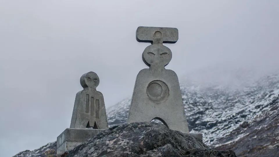To moderne stenskulpturer med abstrakte menneskelignende former står på en klippe i et snedækket, bjergrigt landskab under en grå himmel.