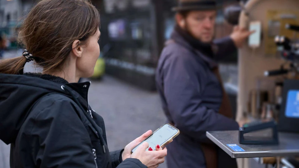 Kvinde betaler med mobiltelefon ved en udendørs kaffevogn, hvor en barista forbereder en bestilling.