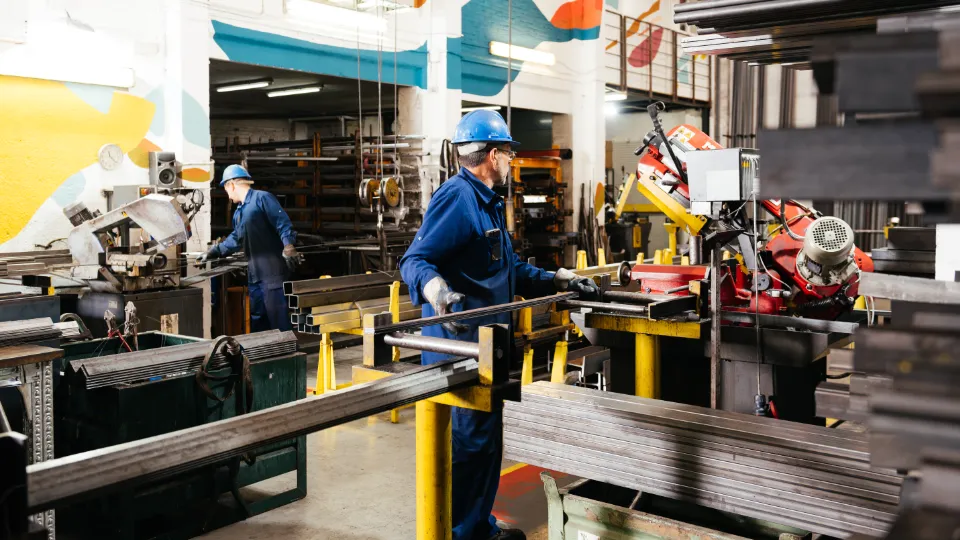 To industriarbeidere med verneutstyr jobber med metallbearbeiding i en fabrikkhall omgitt av maskiner og stålrør.