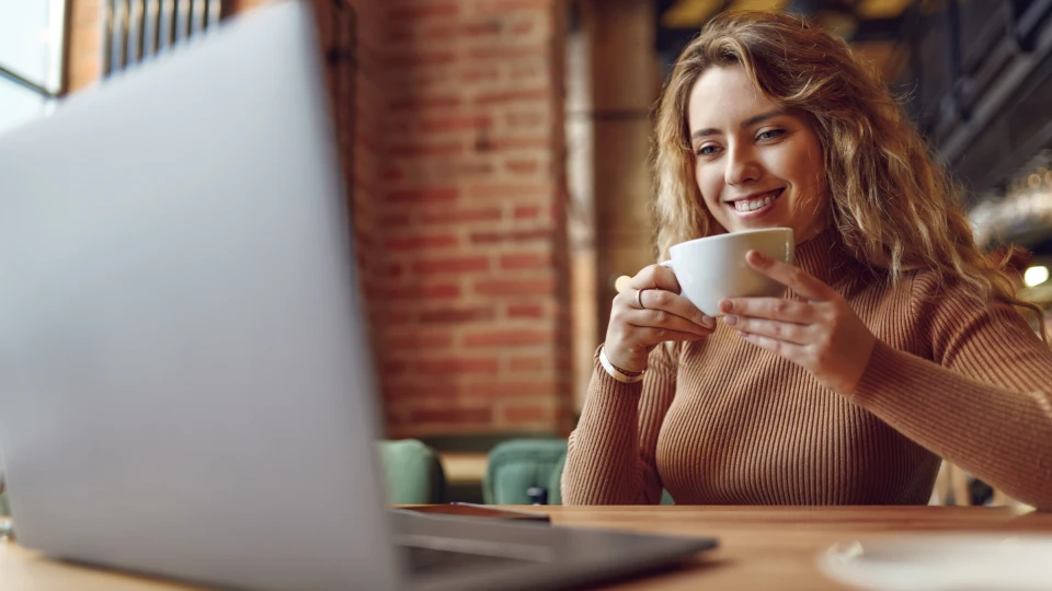Smiling woman in a brown sweater drinking coffee while looking at a laptop in a cozy café.