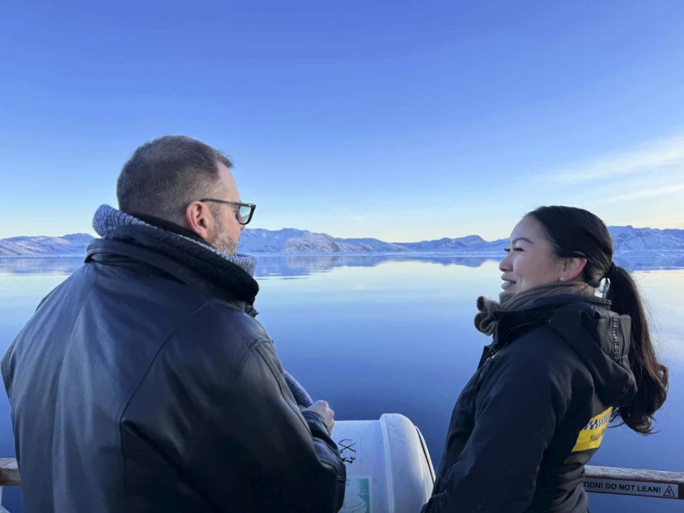 Tristram Hooley og Pakkutannguaq Larsen står på en båd i Grønland og samtaler med snedækkede bjerge og spejlblankt vand i baggrunden.