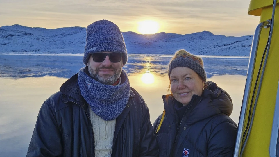 Tristram Hooley og Rie Thomsen på en båd i Grønland med sneklædte bjerge og solnedgang i baggrunden.