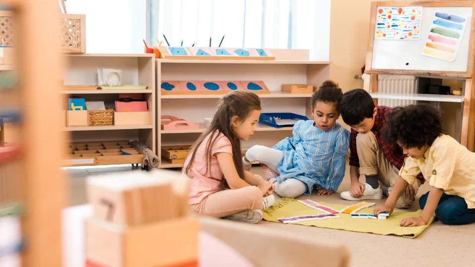 Fyra barn i förskoleåldern sitter tillsammans på ett ljust golv och leker med färgglada pedagogiska verktyg. De fokuserar på en aktivitet med olikfärgade brickor som ligger utspridda på en liten matta. I bakgrunden syns ljusa trämöbler med prydligt sorterat material och en whiteboard, vilket skapar en inspirerande läromiljö.