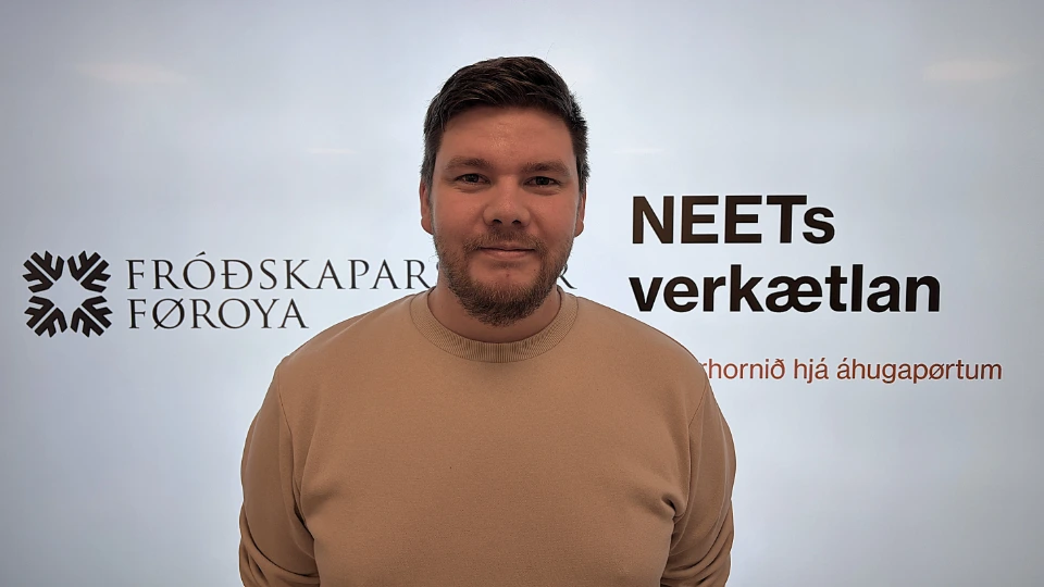 Hallur Mohr Sverrason, der står foran en hvid baggrund med tekst og logoer. Han har mørkt hår, kort skæg og er iført en beige sweatshirt. Bag ham ses logoet for Fróðskaparsetur Føroya og teksten "NEETs verkætlan"