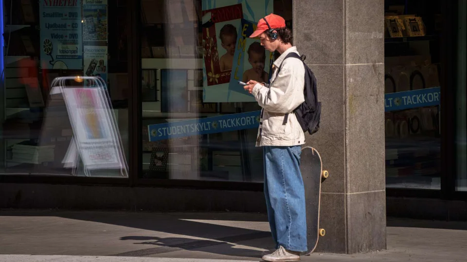 Ung mand med skateboard står ved en butik og kigger på sin telefon, iført rød kasket, høretelefoner og rygsæk.