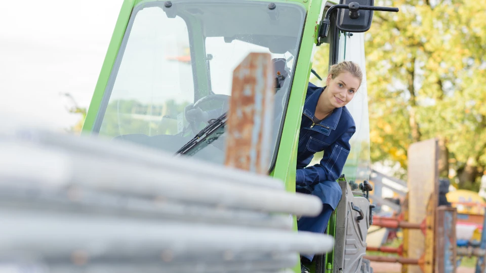En smilende ung kvinne i blå arbeidsdress sitter i førerhuset på et lysegrønt anleggskjøretøy eller en gaffeltruck. Hun ser ut mot kameraet mens hun sitter ved rattet. I forgrunnen er det noen uskarpe metallrør, og i bakgrunnen kan man se trær med grønt løv.
