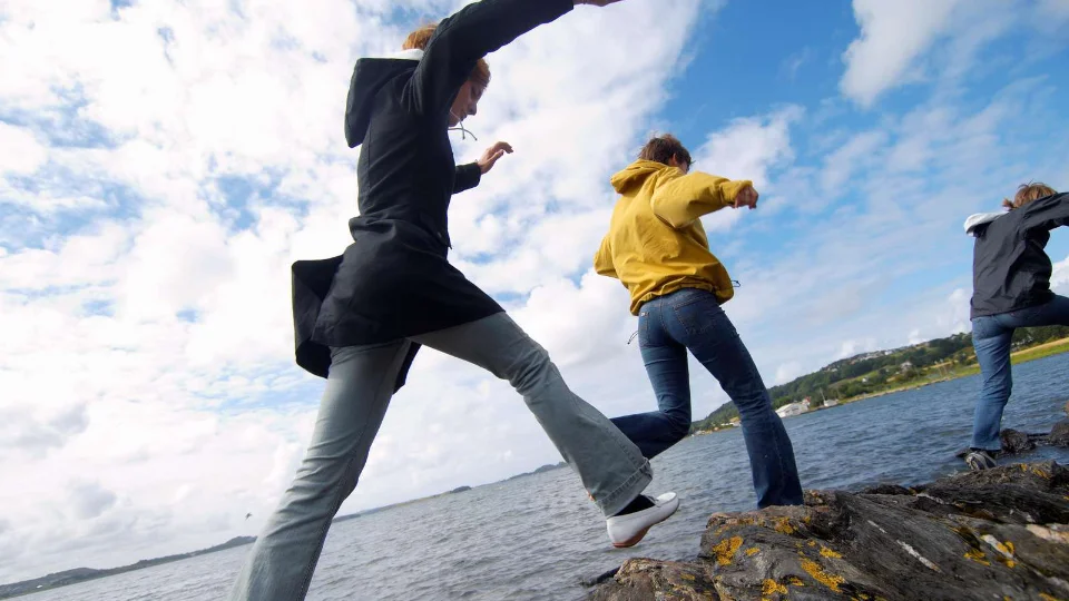 Tre unga personer hoppar lekfullt mellan klippor vid ett utbrett vatten under en blå himmel med vita moln. Bilden är tagen ur ett lågt perspektiv som betonar rörelsen och känslan av frihet.