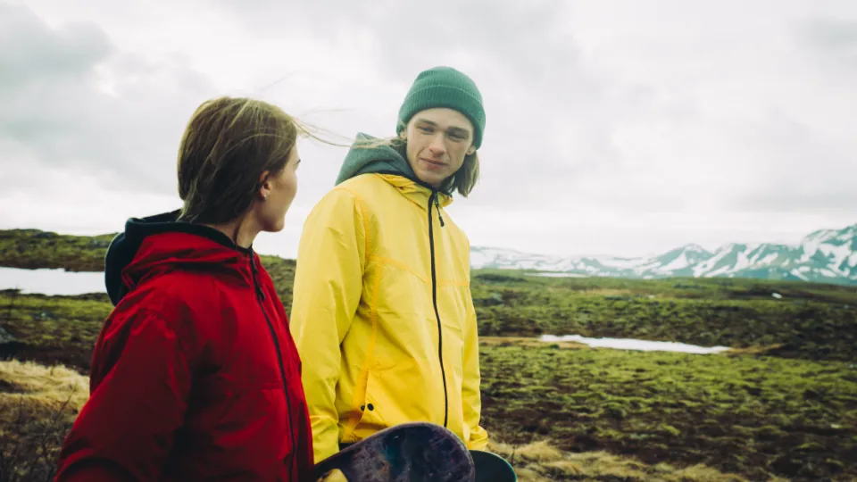 Hvorfor får så få grønlandske unge en uddannelse? To personer i farverige jakker står i et åbent landskab med mos, søer og snedækkede bjerge i baggrunden.