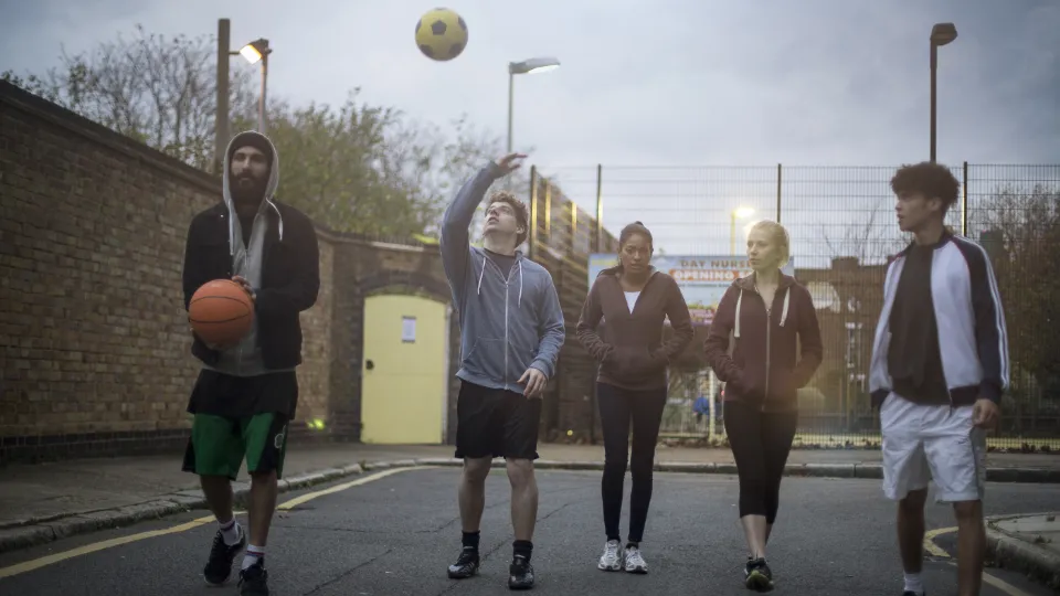 Forebyggende arbeid for barn og unge styrkes med over en halv milliard Fem unge voksne går på en urban gate med basketball og fotball ved en inngjerdet bane.