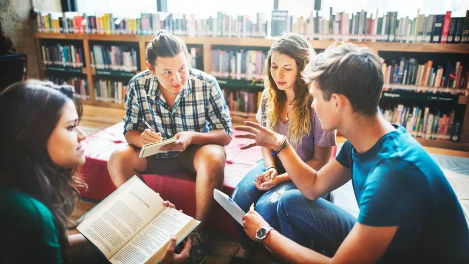 Fire unge sidder i en rundkreds på et bibliotek og diskuterer, mens én læser i en bog og en anden tager noter.